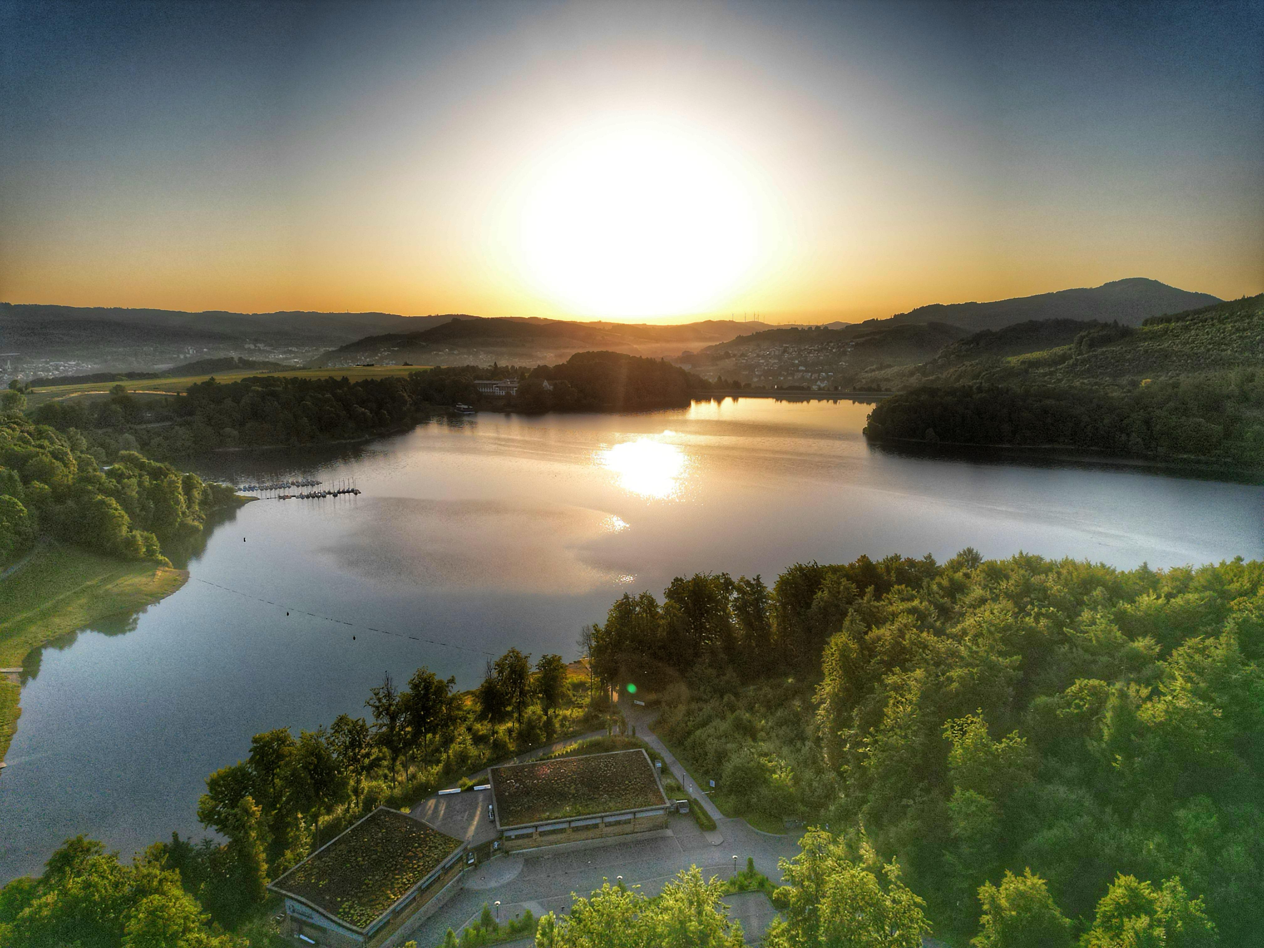 Mit dem H1 am See eröffnete 2018 eine Eventlocation der Extraklasse am Hennesee bei Meschede Ein See, umgeben von Bäumen und Gebäuden.
